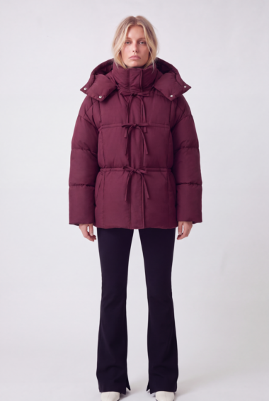 Woman wearing a burgundy puffer coat with tied toggles and a hood, standing against a light background.
