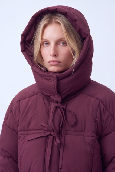 Woman wearing a maroon puffer jacket with a large hood, facing the camera, neutral expression.