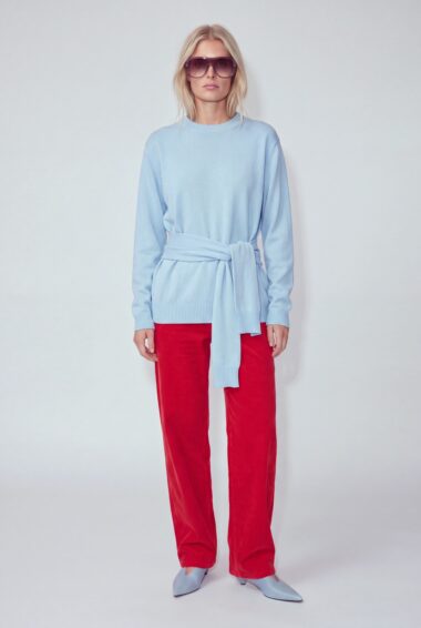 Full-body portrait of a woman in a light blue belted sweater, bright red pants, and lavender sunglasses against a pale backdrop.