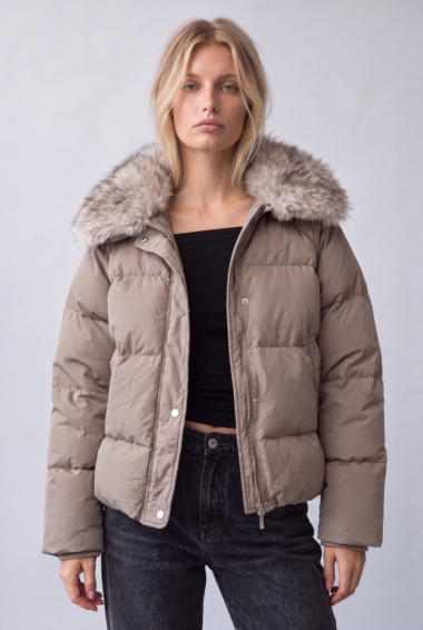 Woman wearing a taupe padded jacket with a fluffy fur-trimmed hood, black top, and dark jeans, posing against a light gray backdrop.