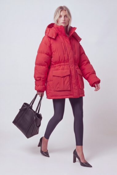 Woman in a bright red oversized puffer coat with a high collar, black leggings, and black heeled pumps walks while carrying a dark handbag in a studio setting.